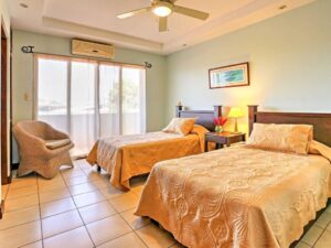 Bright guest bedroom with two twin beds, air conditioning, built-in closets, large window with greenery view, and en-suite bathroom.